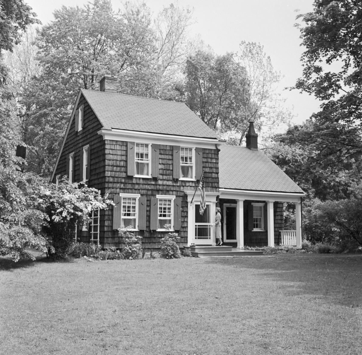 The Thomas Paine Cottage photographed from its south lawn in about 1950: a shingled colonial house with dark shutters, a small covered porch, and heavy trees behind it.