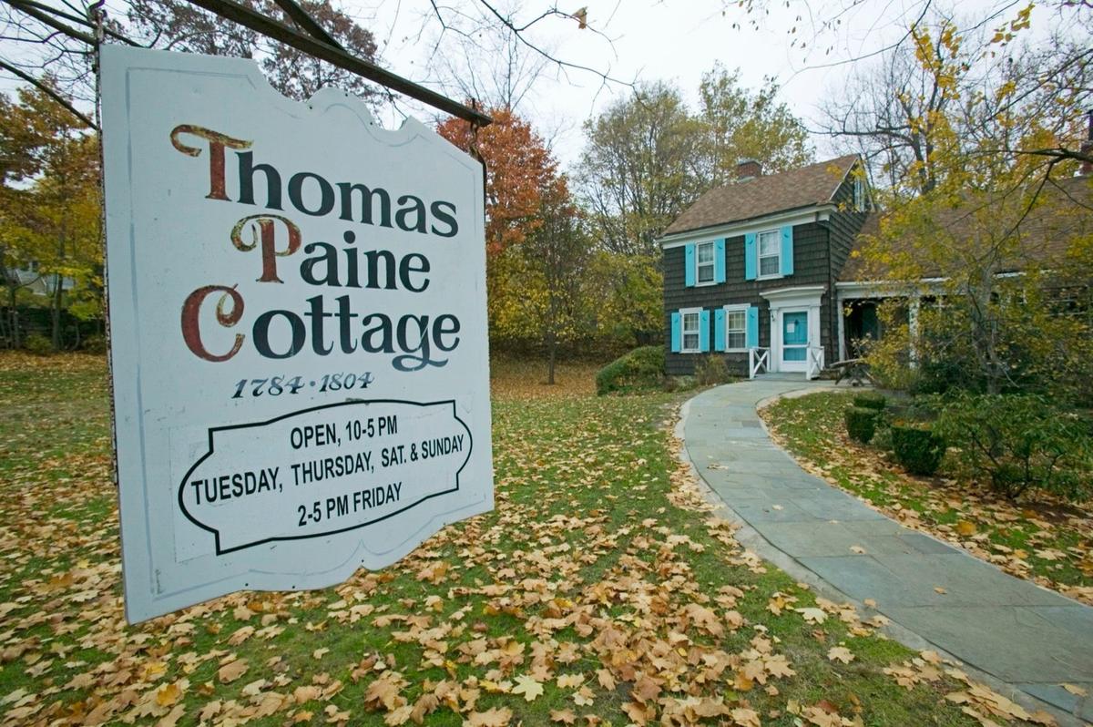 A painted wooden sign reading Thomas Paine Cottage, 1784-1804, with visiting hours, standing in deep fallen leaves. The dark shingled cottage sits at the end of a path behind it.