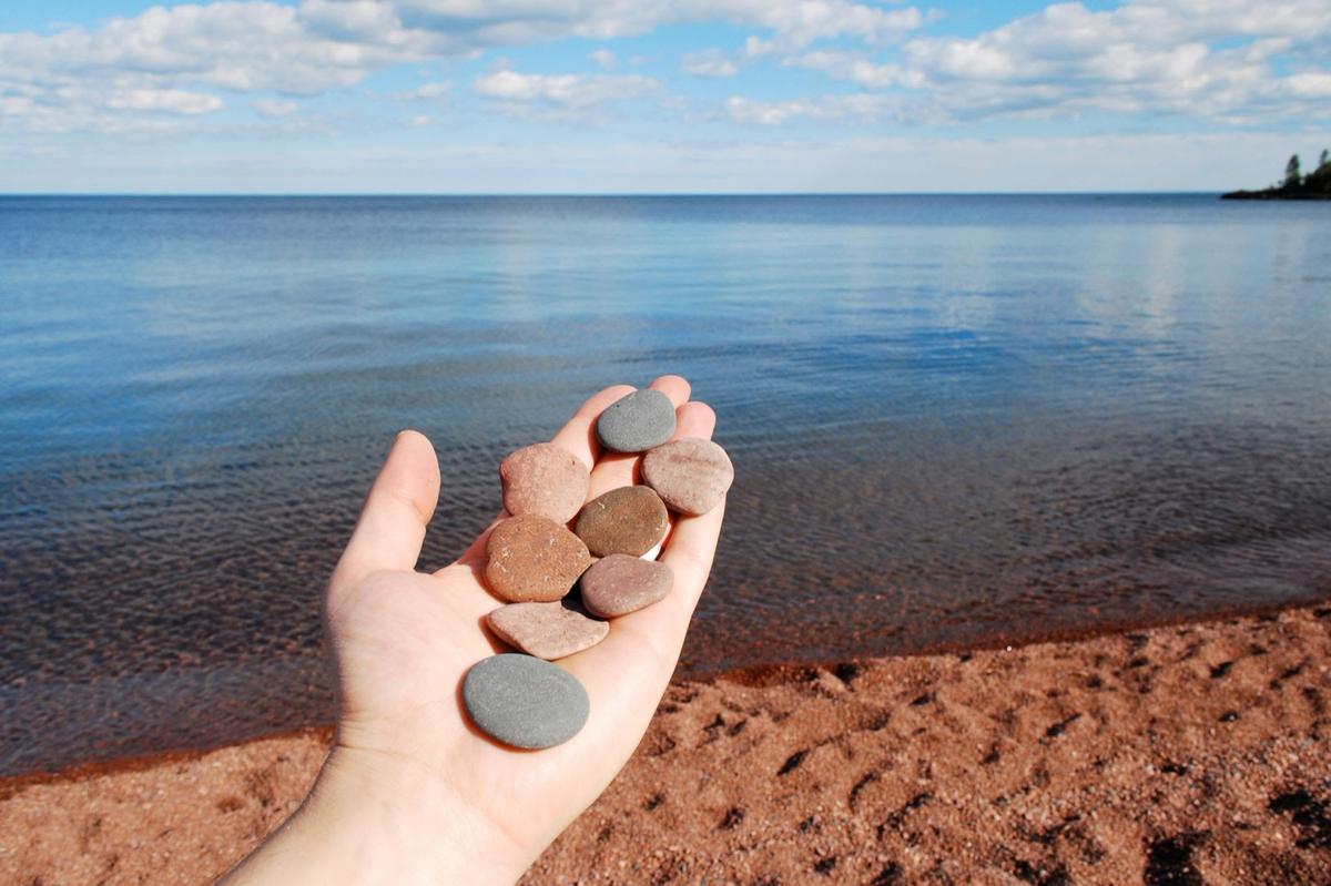 A hand holding a palmful of smooth flat skipping stones above a shingle beach, with flat open water running to the horizon.