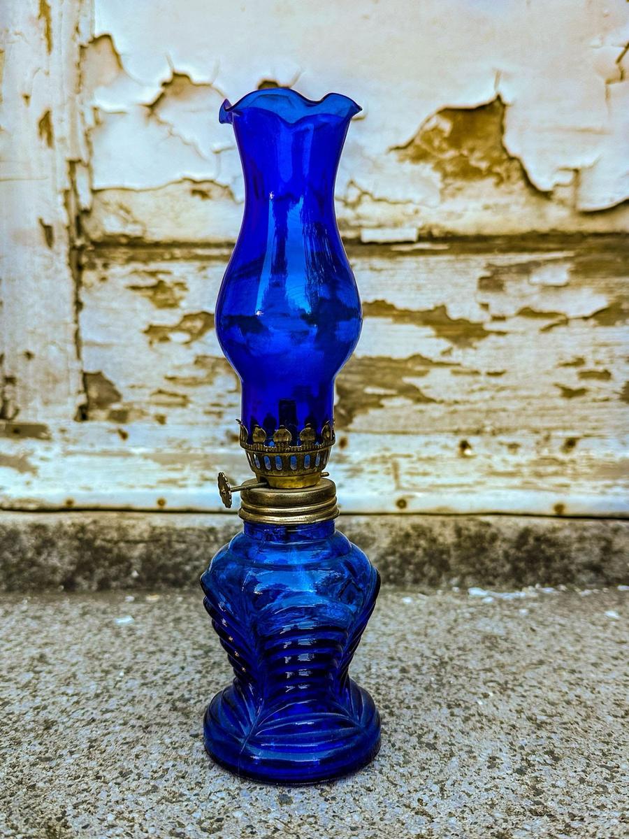 A small cobalt-blue pressed glass oil lamp with a fluted chimney and brass collar, standing against a weathered white door.